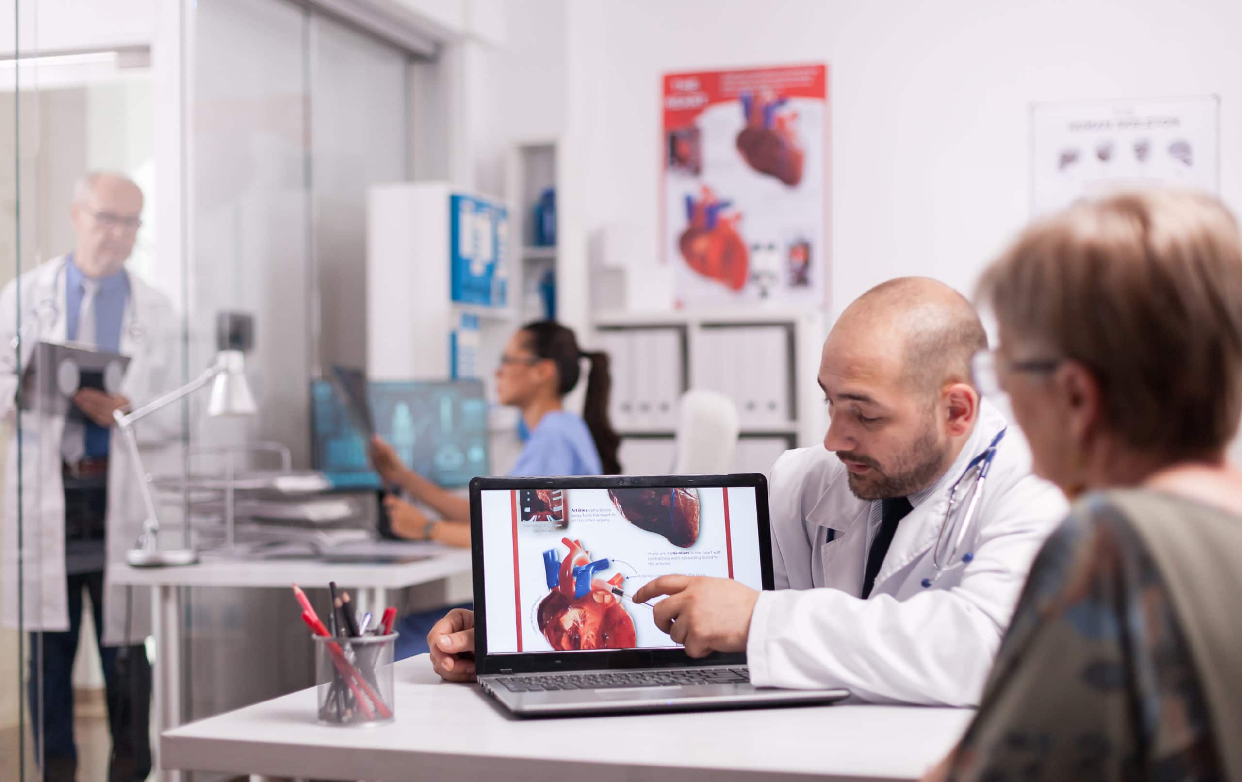 Young doctor pointing at heart on laptop screen in hospital office. Cardiologist with senior patient during consultation. Mature medic taking notes on clipboard on clinic corridor.