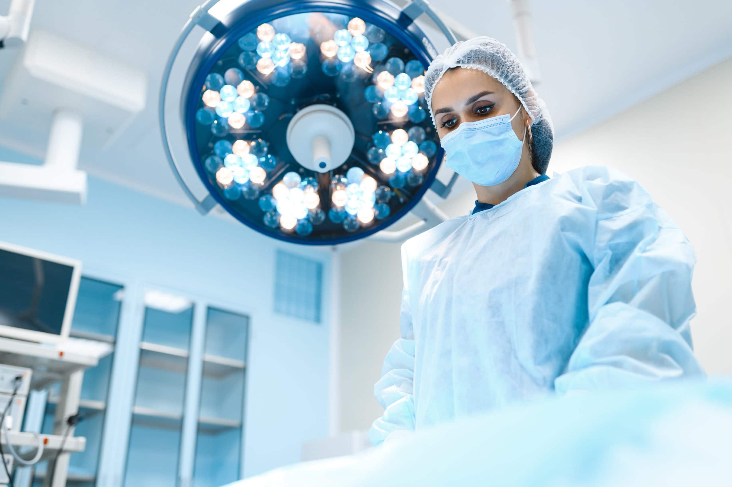 Female surgeon in gown and mask in operating room, lamp on background, surgery operation. Doctor in uniform, medical clinic worker, medicine and health, healthcare in hospital